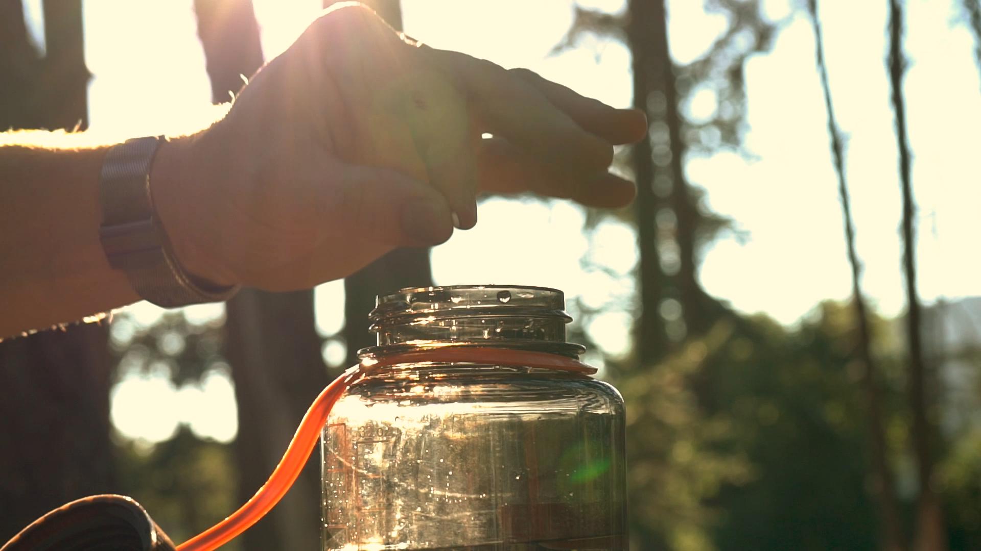 Close-up of hand preparing to treat water in a reusable bottle in a sunlit forest; water purification is crucial for waterborne illness prevention outdoors.