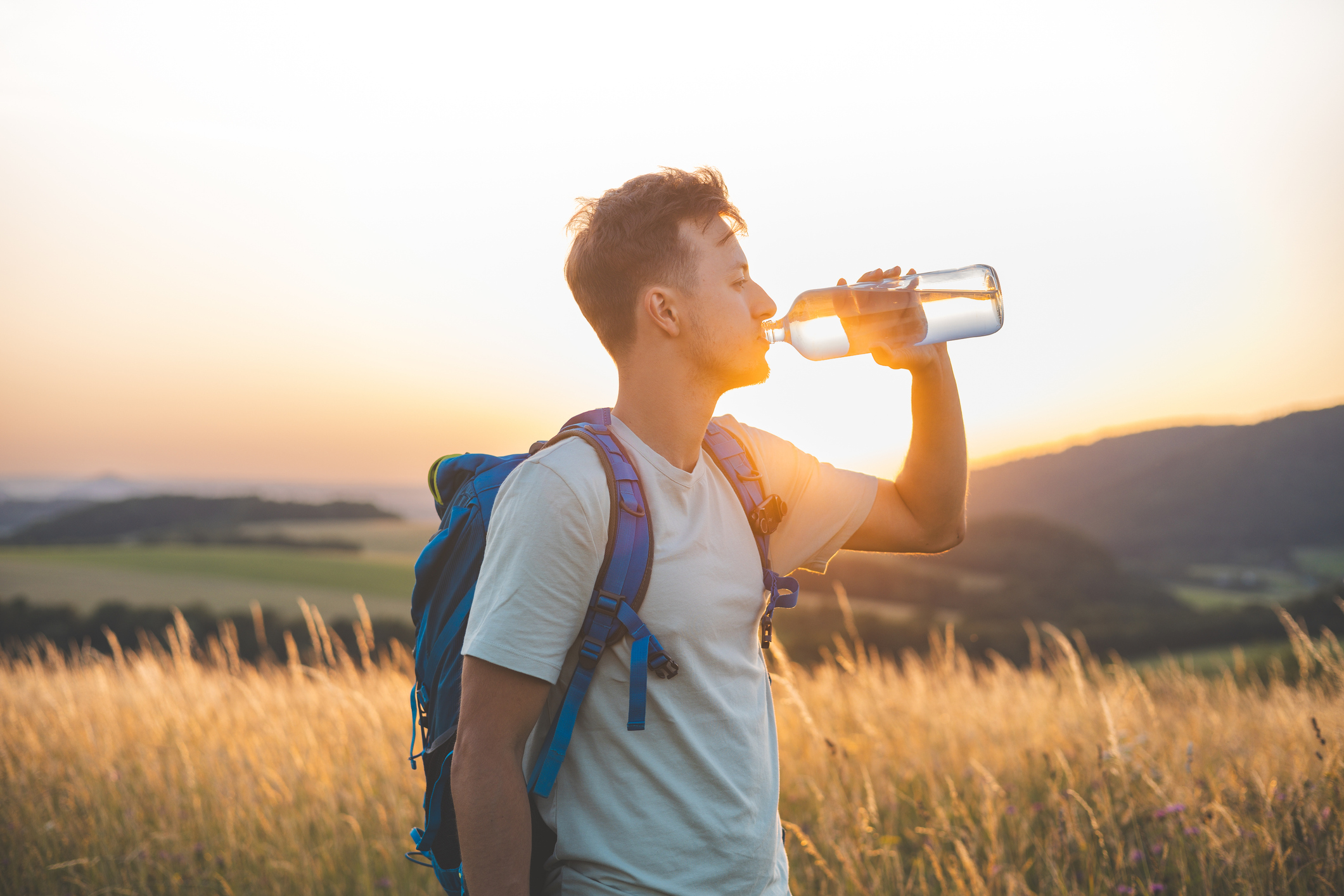 Young man with backpack hydrates from a water bottle while hiking in a field at sunset. Access to safe water outdoors is key for waterborne illness prevention.