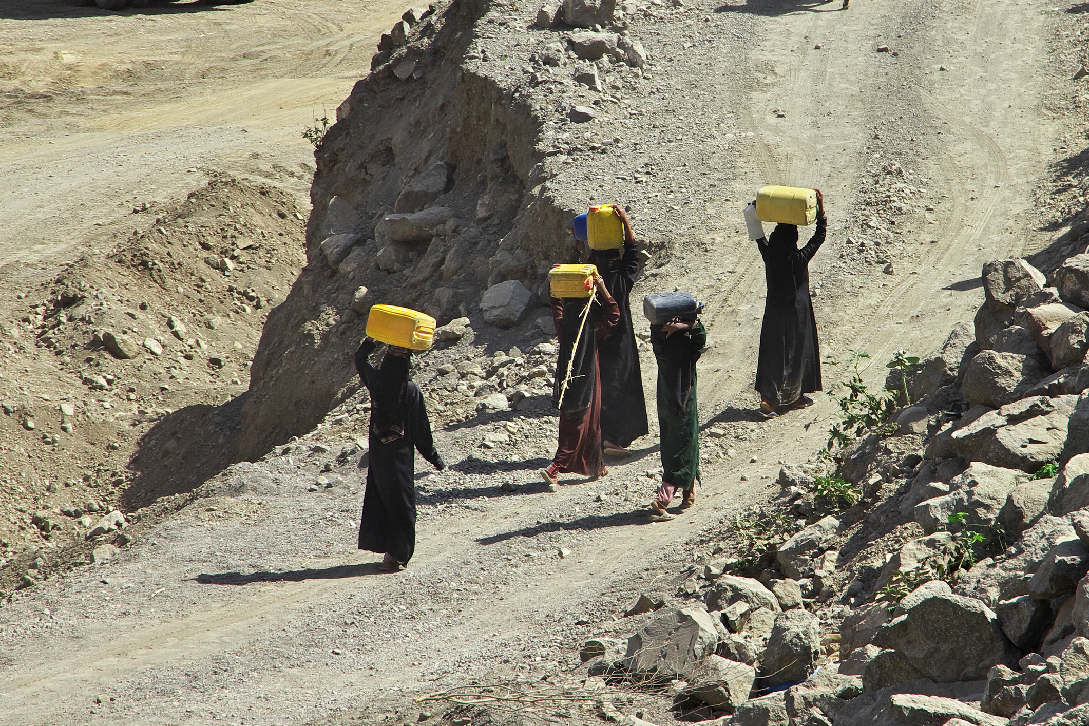 People walking on a rugged path carrying water containers, depicting the challenge of accessing water in difficult terrain.